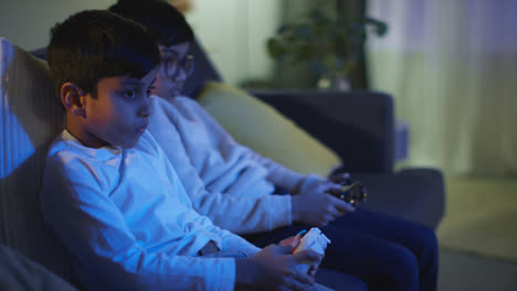 Side-View-Of-Two-Young-Boys-At-Home-Having-Fun-Playing-With-Computer-Games-Console-On-TV-Holding-Controllers-Late-At-Night